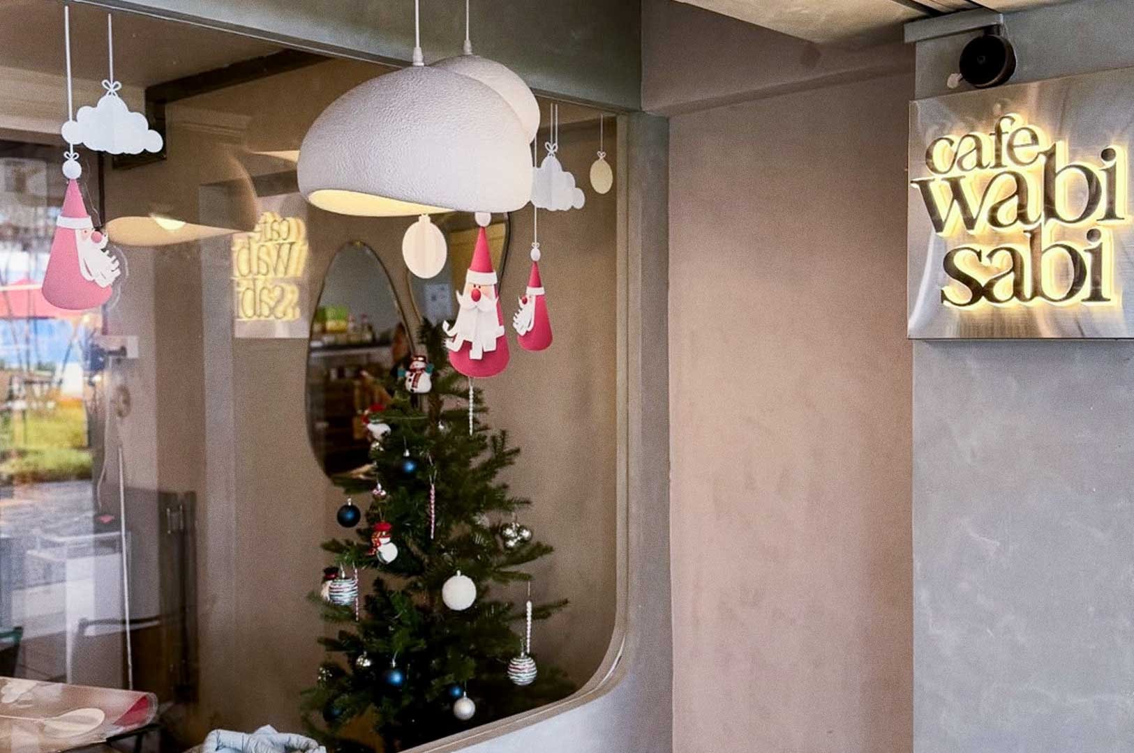Exterior shot of a cafe wabi sabi window dressed for the holidays, featuring a glowing name sign and interior Christmas tree with ornaments and a small snowman figure.