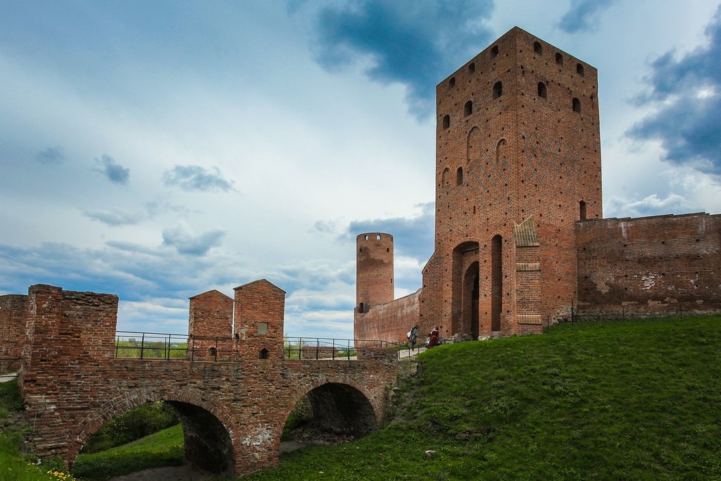 Zamek, a właściwie jego ruiny, o gotyckiej architekturze z wysoką, ceglano-murarską wieżą i mostem łukowym na zielonym terenie. Widok zamku otoczonego murami obronnymi i niebem pełnym chmur. (źródło: https://www.flickr.com/photos/polandmfa/26231981854)