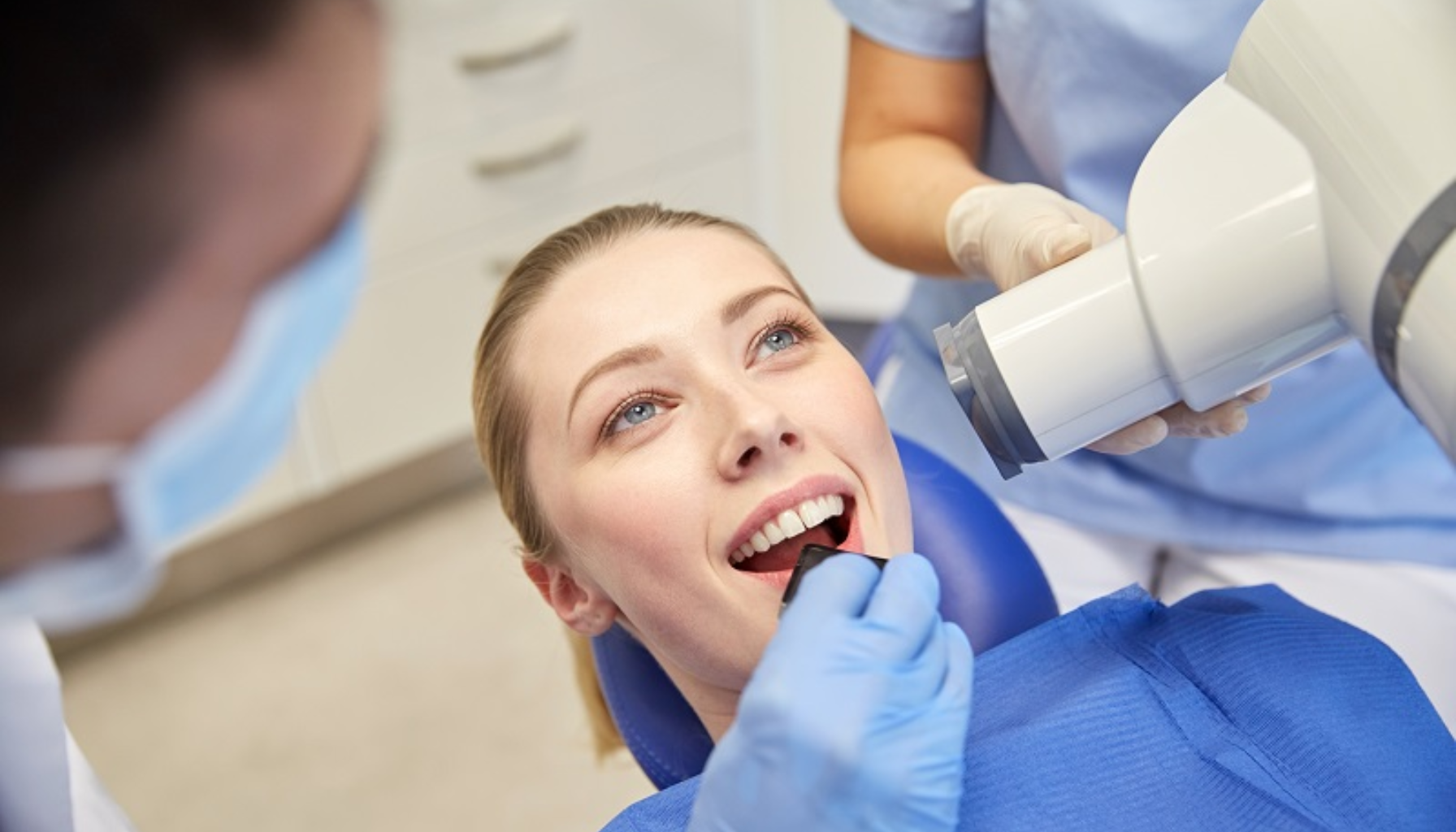 Kvinne hos tannlegen får intraoralt røntgen, holder sensor i munnen mens helsepersonell justerer røntgenapparatet og gir instruksjoner.