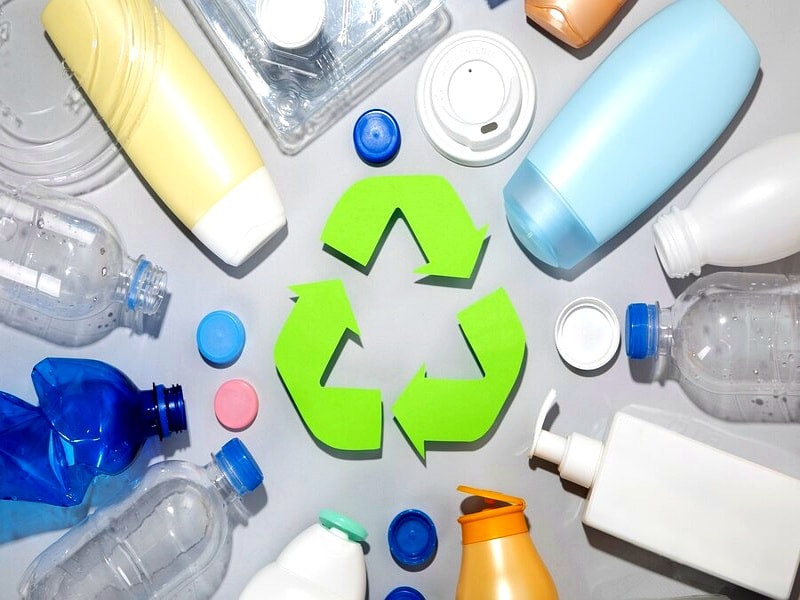A group of plastic bottles, caps, and containers arranged in a circle around a green recycling symbol on a light background.