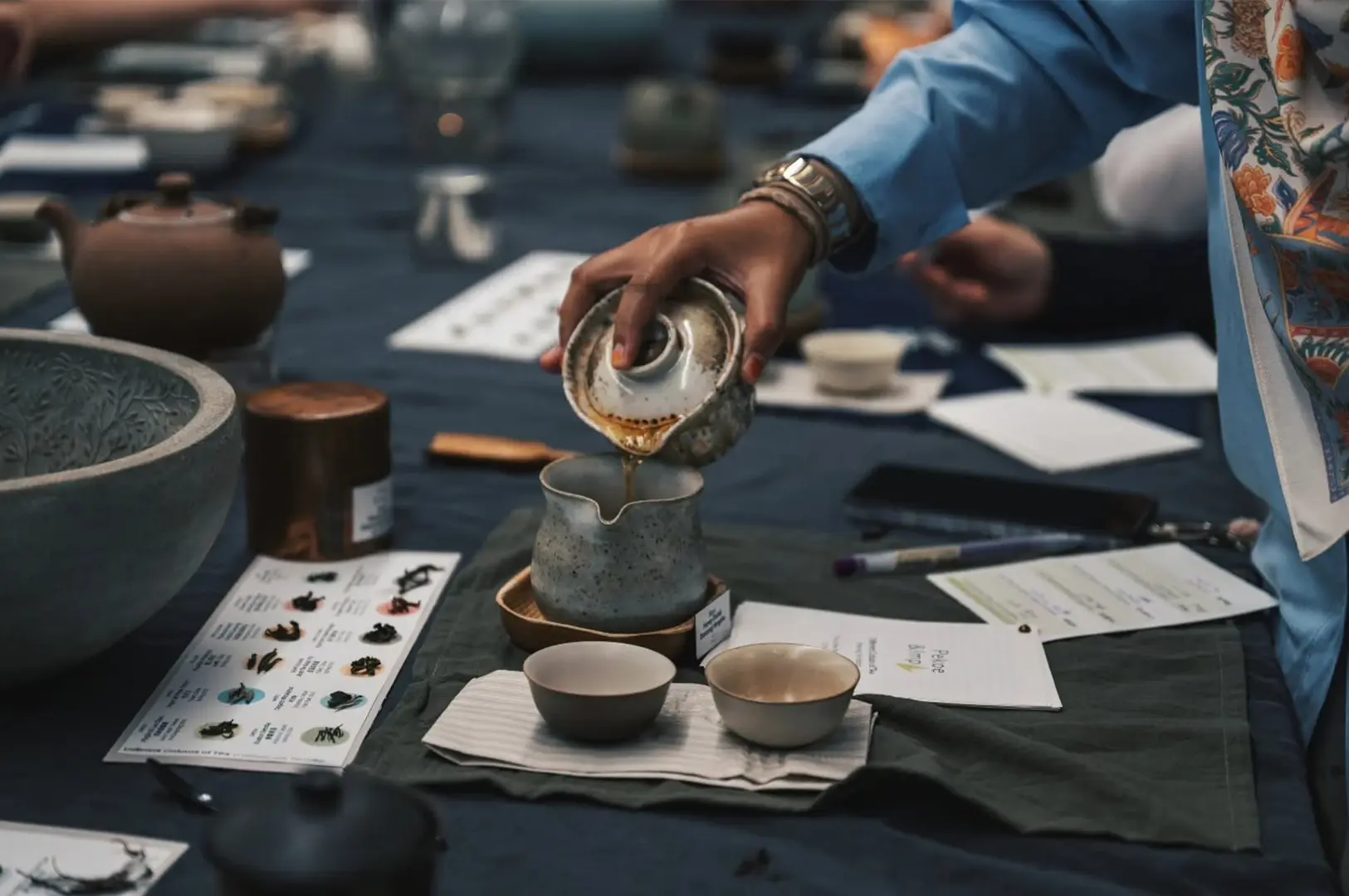 An eye-level shot of a person's hand pouring hot tea from a rustic, dark-colored clay pitcher into one of several small white cups arranged on a dark table. Steam rises elegantly from the cups and the pouring stream, creating a soft, atmospheric effect against the diffused background.