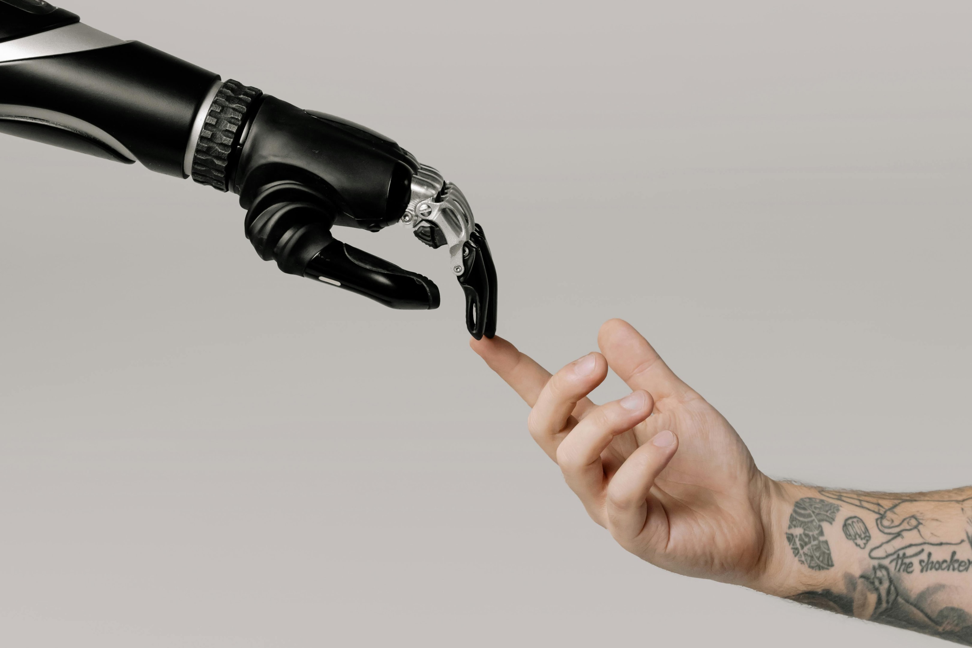 "Close-up image of a human hand with tattoos reaching out to touch the finger of a robotic hand, symbolizing the connection between humans and technology. The background is a neutral gray, emphasizing the interaction and contrast between organic and artificial intelligence