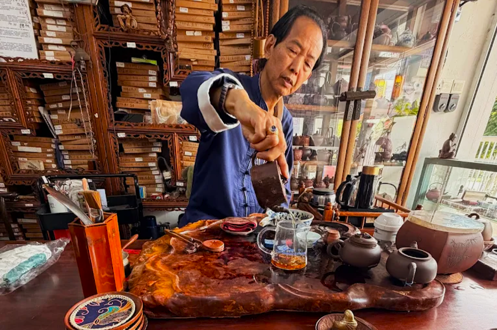An elderly man in a blue shirt pours tea from a pot at a cluttered wooden table in a cozy, traditional tea shop. The setting exudes warmth and tranquility.