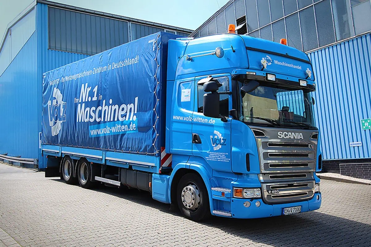 Blauer Lastwagen der Firma Volz, geparkt vor einer Lagerhalle. Der Lkw trägt die Aufschrift 'Nr. 1 in Maschinen!' und die Website-Adresse 'www.volz-witten.de'. Das Fahrzeug dient dem Transport von Maschinen und vermittelt Professionalität im Maschinenhandel.