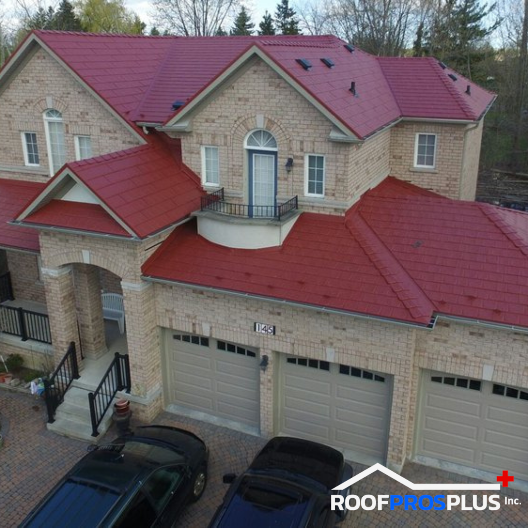 A two-story brick house with bold red metal roofing, featuring arched windows and a small balcony. It has a three-car garage and a paved driveway with two cars in the driveway.