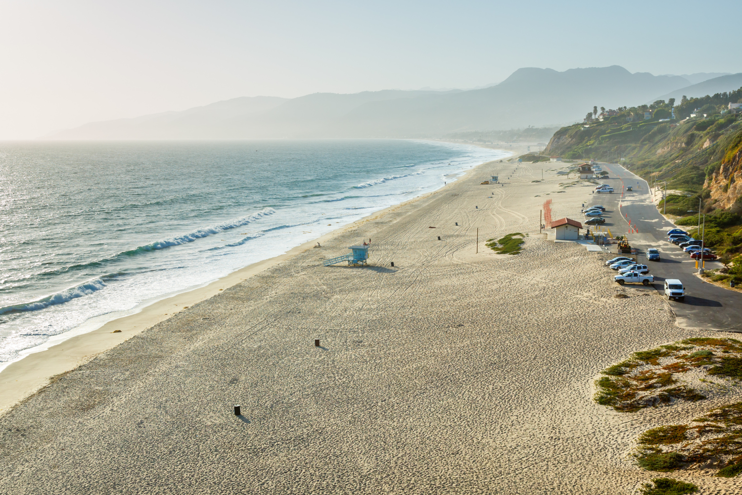 cars parked at road near zuma beach, lifeguard tower at the beach, people and dogs playing