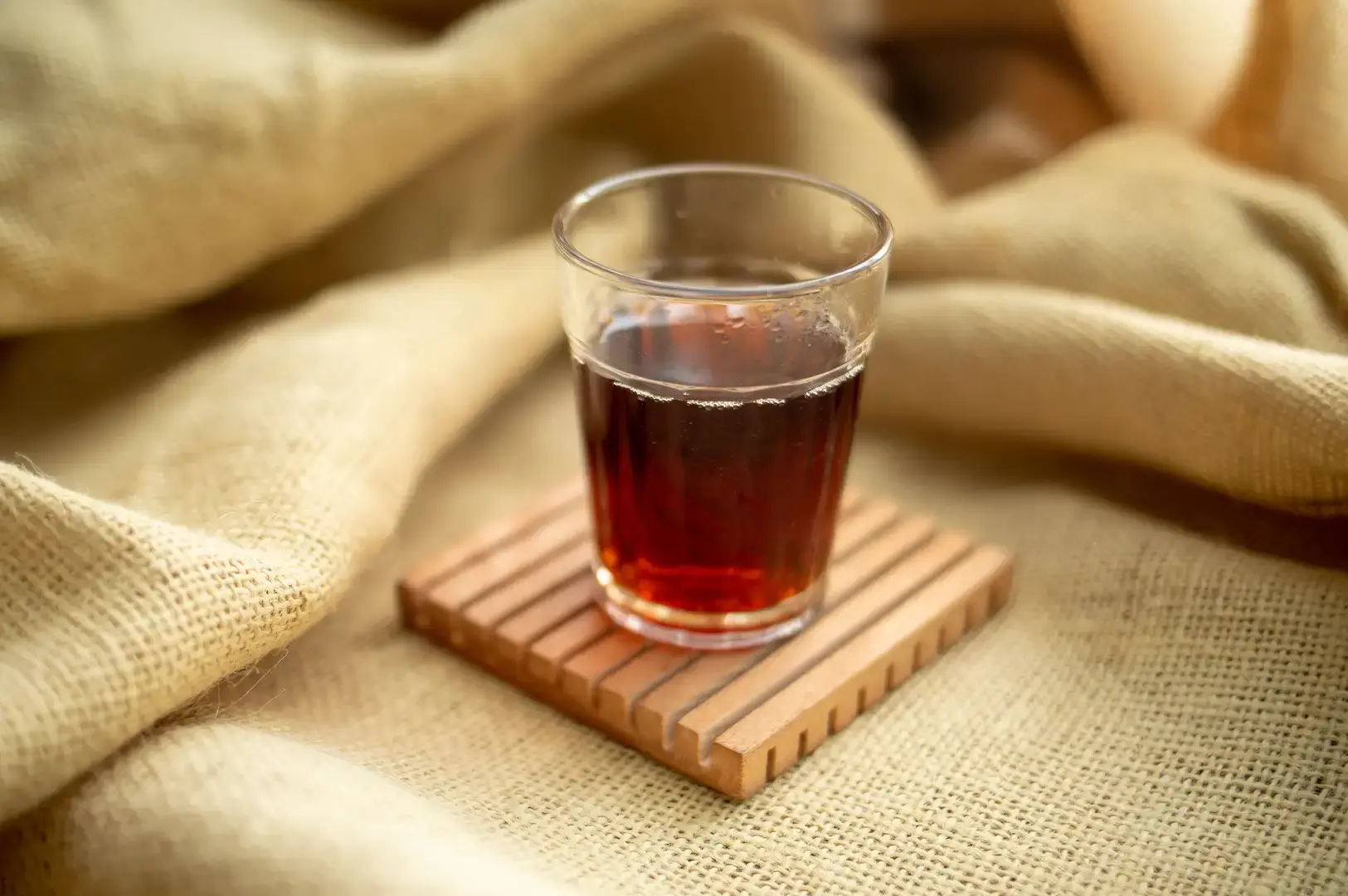 A small, clear glass filled with dark amber tea sits atop a square wooden slotted coaster, which rests on draped, warm beige burlap fabric. The soft focus and warm illumination create a cozy, rustic atmosphere around the minimalist tea setup.