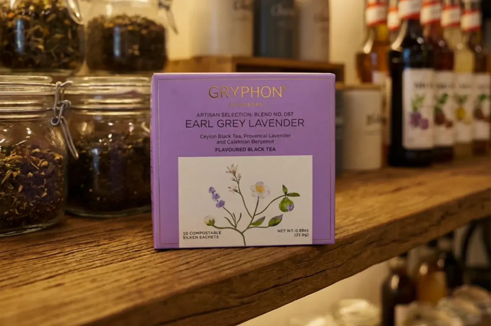 A lavender-colored square box of Gryphon Singapore's Artisan Selection Blend No. 087, "Earl Grey Lavender," stands centered on a rustic, dark-toned wooden shelf. The box design includes elegant gold lettering and a white lower section illustrated with a delicate sprig of lavender and white blossoms, noting that it contains 10 compostable silken sachets of flavoured black tea. The background shows a cozy, shallow-focus cafe or pantry setting with large glass jars filled with loose leaf tea on the left, and blurred glass bottles of syrups or juices lined up on the right.