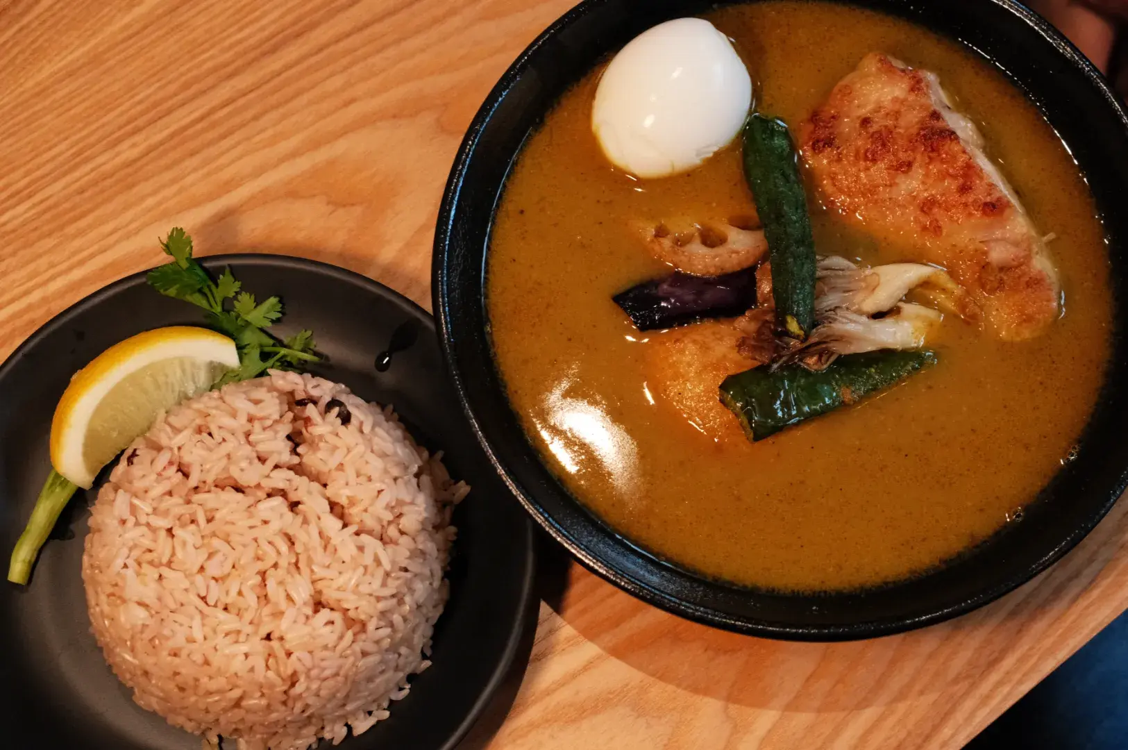 A bowl of soup curry with chicken, vegetables, and a soft-boiled egg, accompanied by a side of brown rice and a lemon wedge on a plate.
