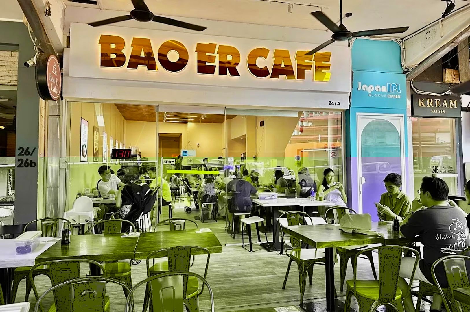 Exterior view of Bao Er Cafe with glass windows revealing a bustling, relaxed atmosphere. Red chairs and wooden tables fill the outdoor seating area.
