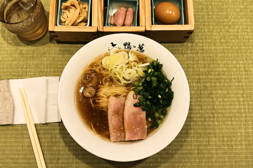 A bowl of ramen with sliced pork, chopped green onions, and leeks in broth, on a green mat. Side dishes include an egg and pickled items.