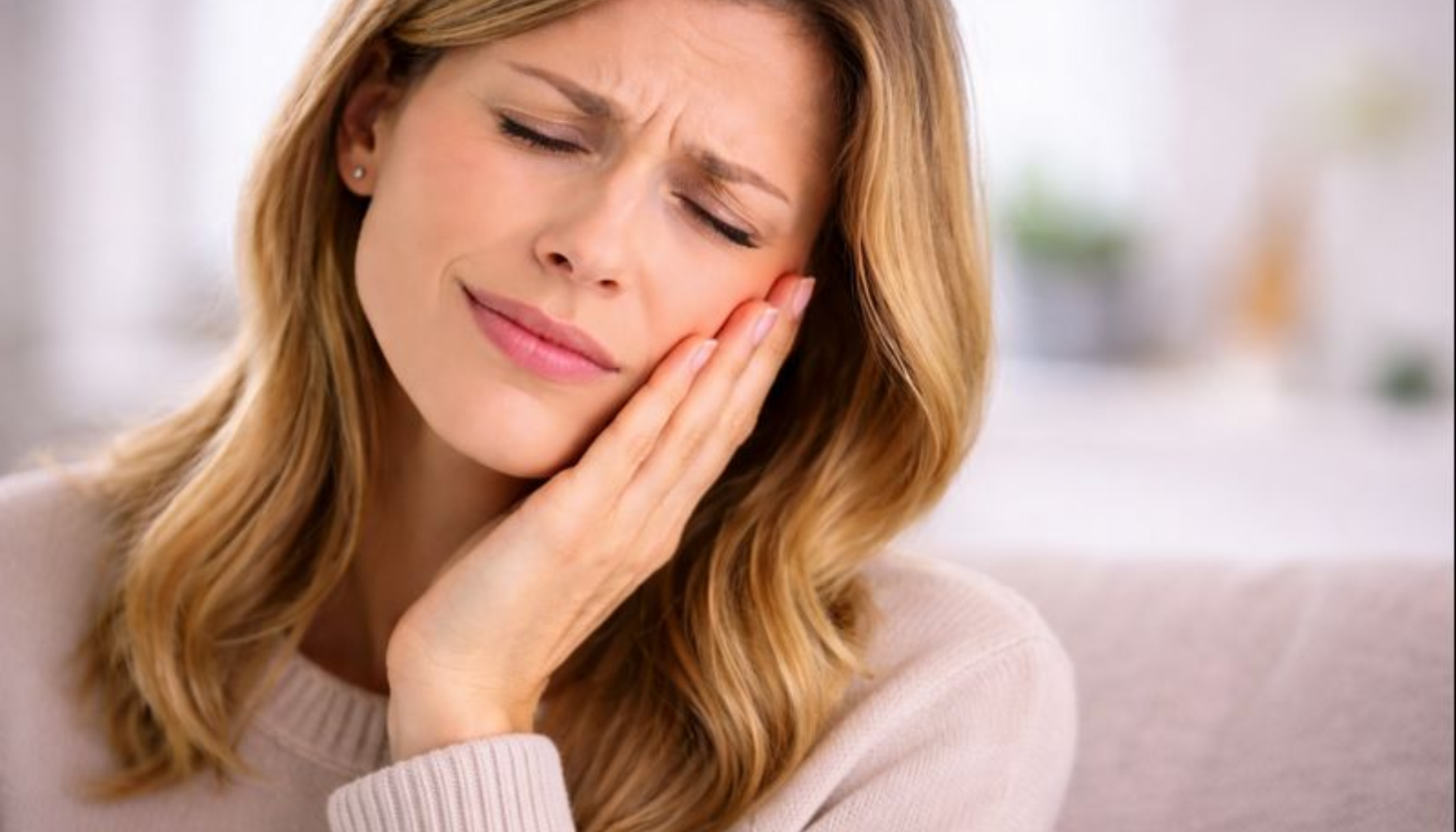 Kvinne holder kinnet forsiktig med mildt ubehag og rolig uttrykk i lyst, hjemlig miljø.