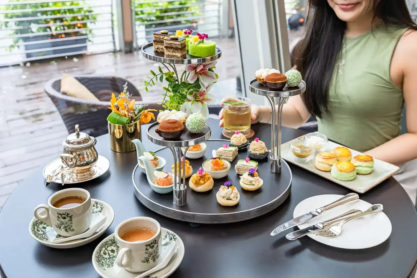 An elegant afternoon tea set is displayed on a dark table, featuring a multi-tiered stand filled with colorful petite cakes, savory bites, and traditional scones. Beside the tiered towers sit vintage-style floral teacups, a silver teapot, and a plate of fresh pastries, all set against a bright, airy patio background.