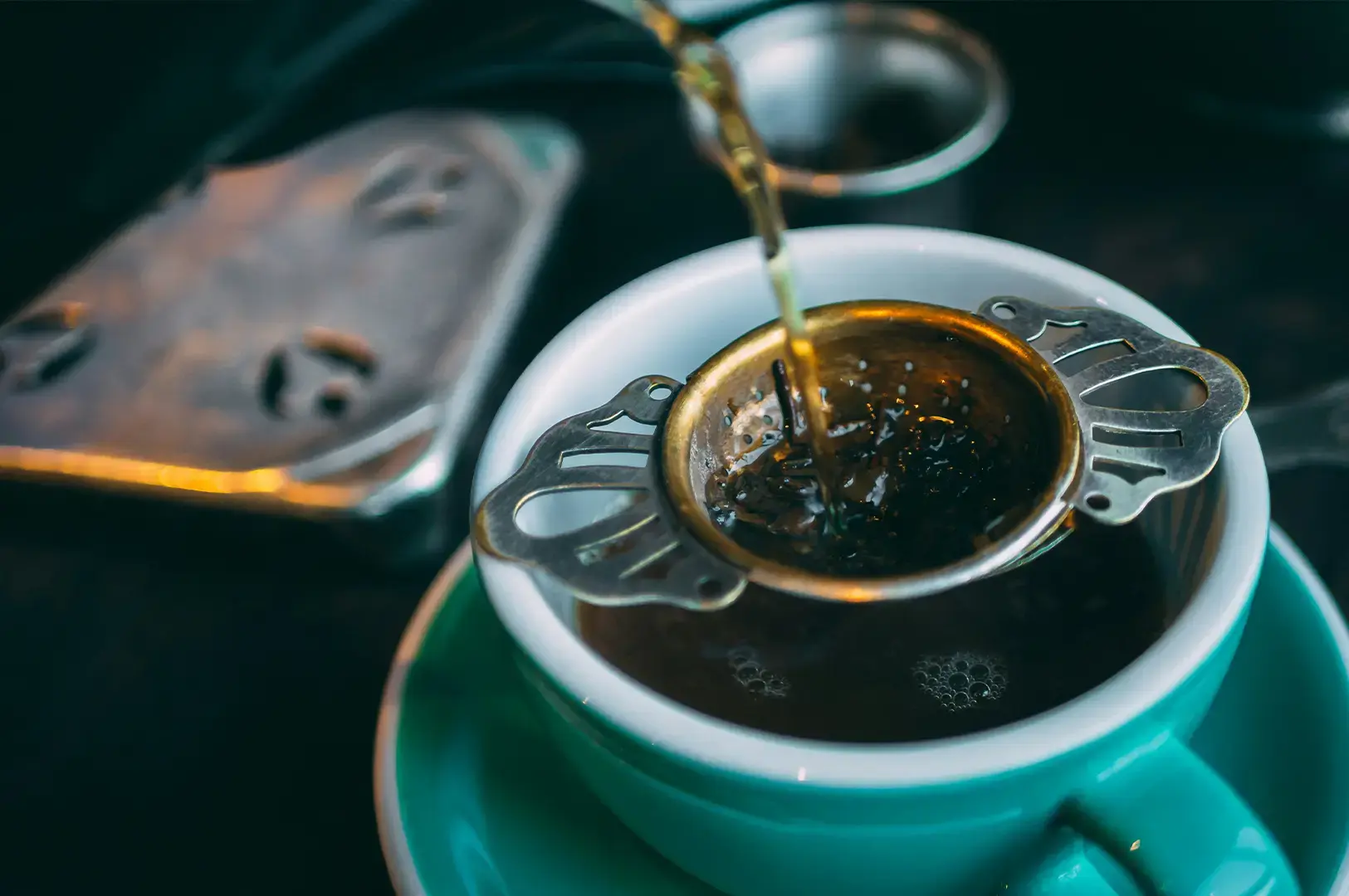 A dramatic close-up captures dark amber tea being poured from above through an ornate, antique-style metal tea strainer balanced over a turquoise teacup. Loose tea leaves are gathered inside the golden-toned bowl of the strainer as the liquid filters down, creating small bubbles on the surface of the steeped tea inside the matching turquoise saucer-supported cup.