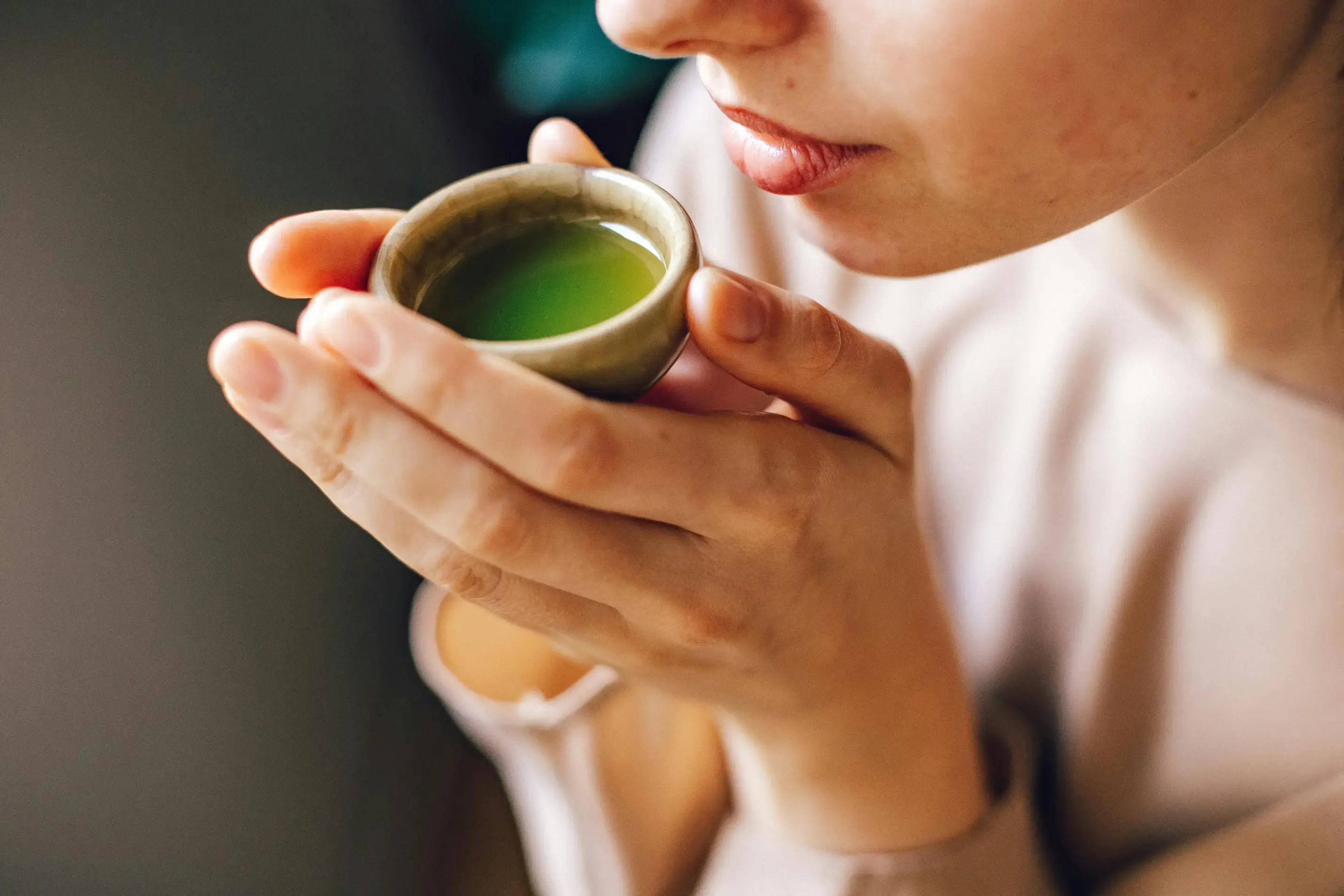 A close-up shot shows a person holding a small, ceramic cup filled with a vibrant green liquid, likely matcha or green tea, up to their lips. Their hands carefully cradle the traditional cup against a soft, neutral-toned background.