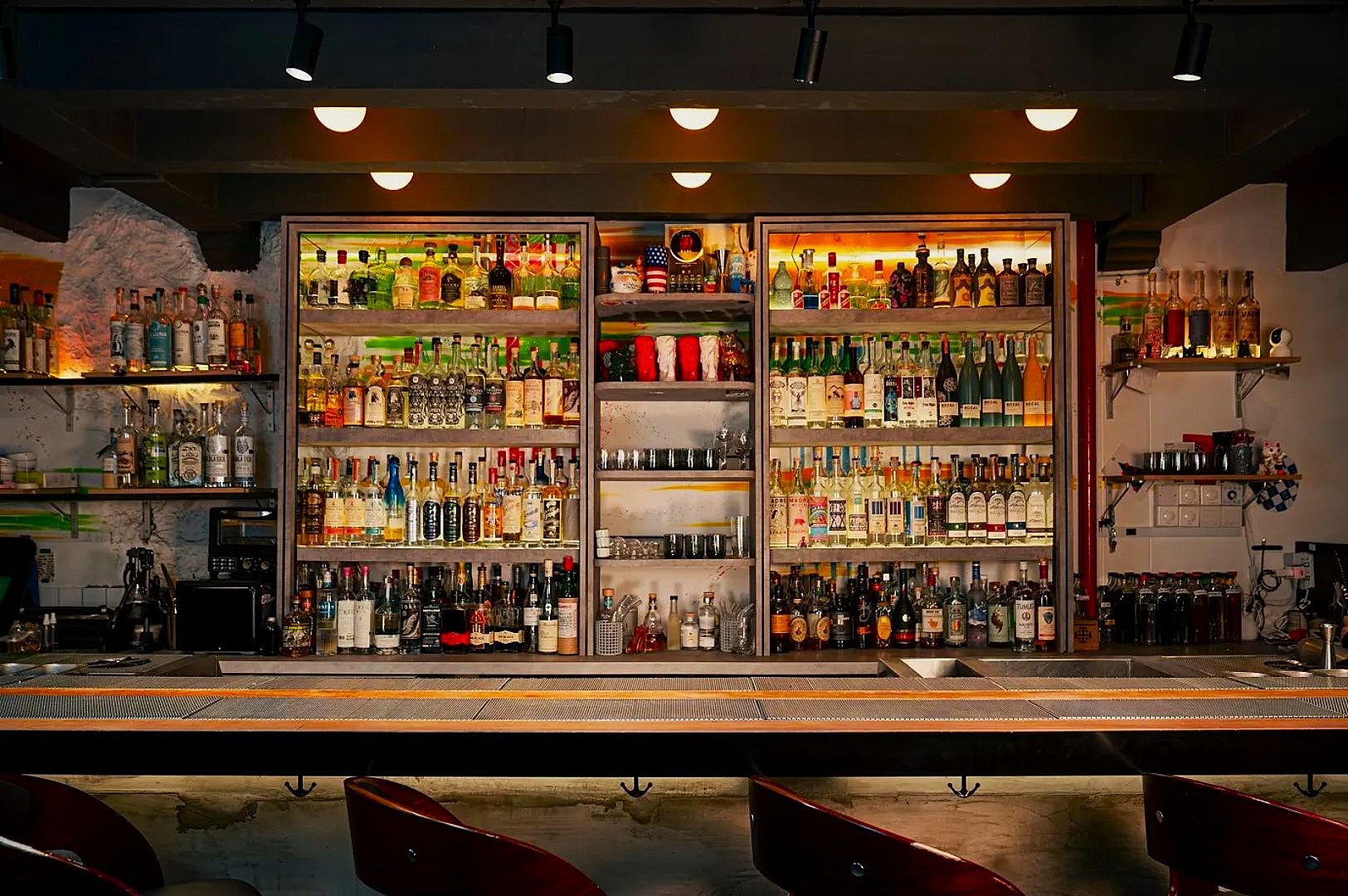 A dimly lit bar showcases a wide array of colorful liquor bottles on backlit shelves behind a sleek wooden counter, creating a warm, inviting atmosphere.