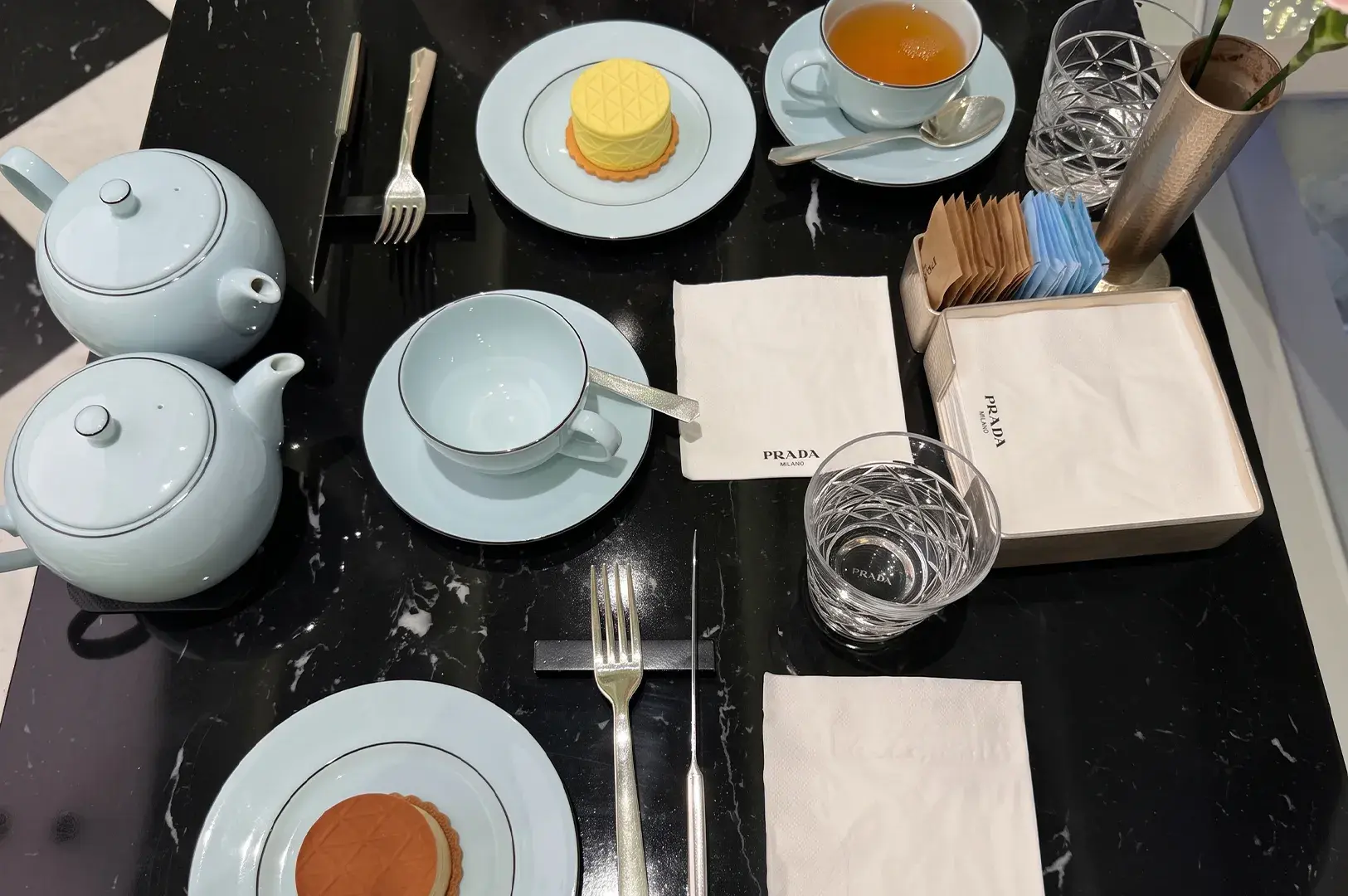 Overhead table layout featuring pale blue teaware, individual dessert plates, teapots, cutlery, and branded Prada paper napkins on black marble.