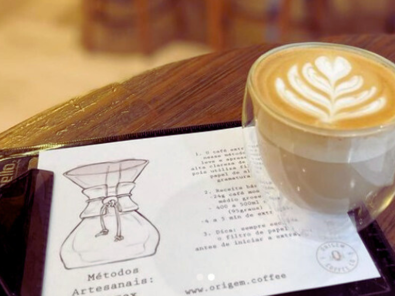 Copo de vidro com latte art sobre folheto explicativo no Origem Coffee CO. Imagem: Reprodução Instagram.