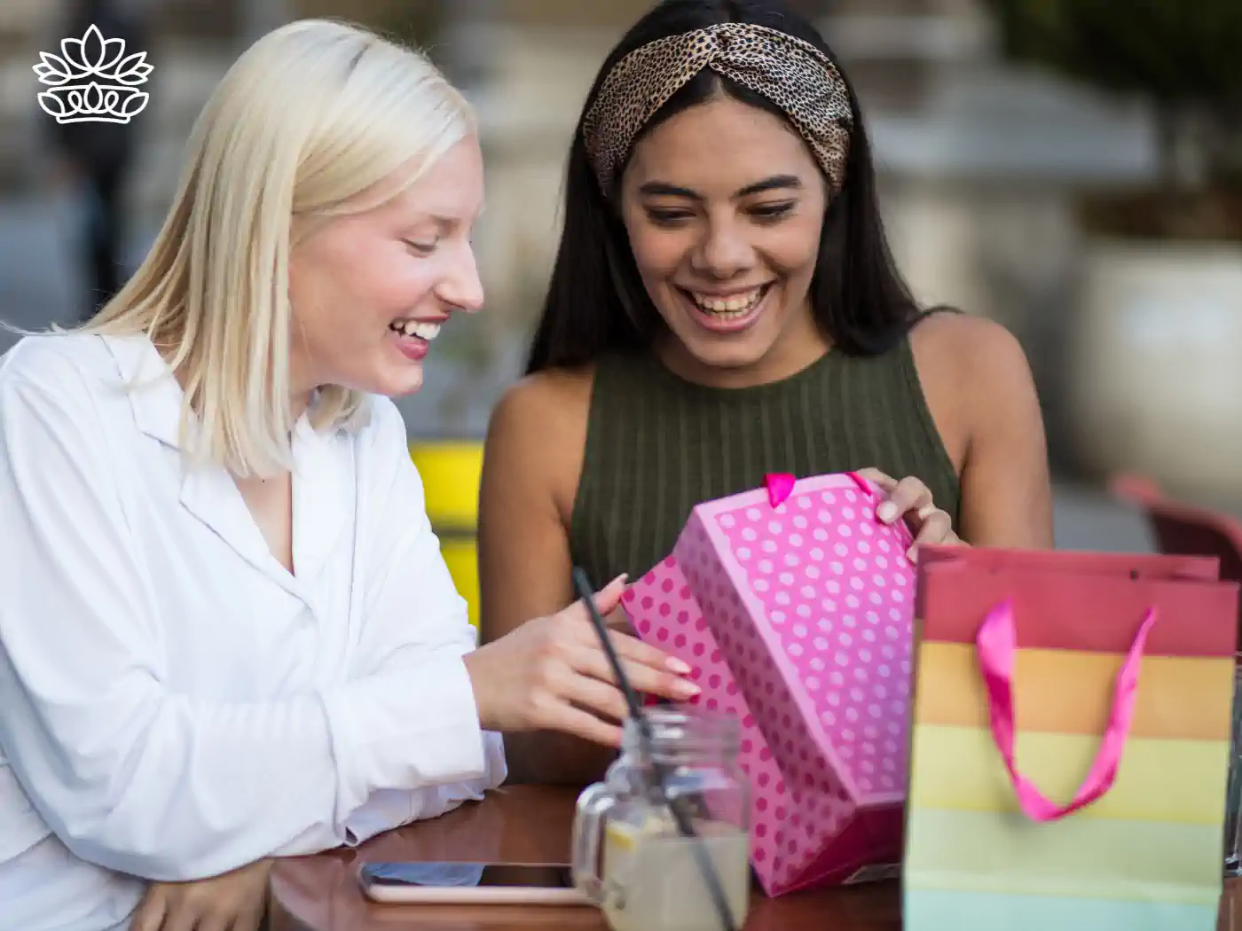 Two women smiling and laughing while opening a pink polka-dotted gift bag at a café. Fabulous Flowers and Gifts.