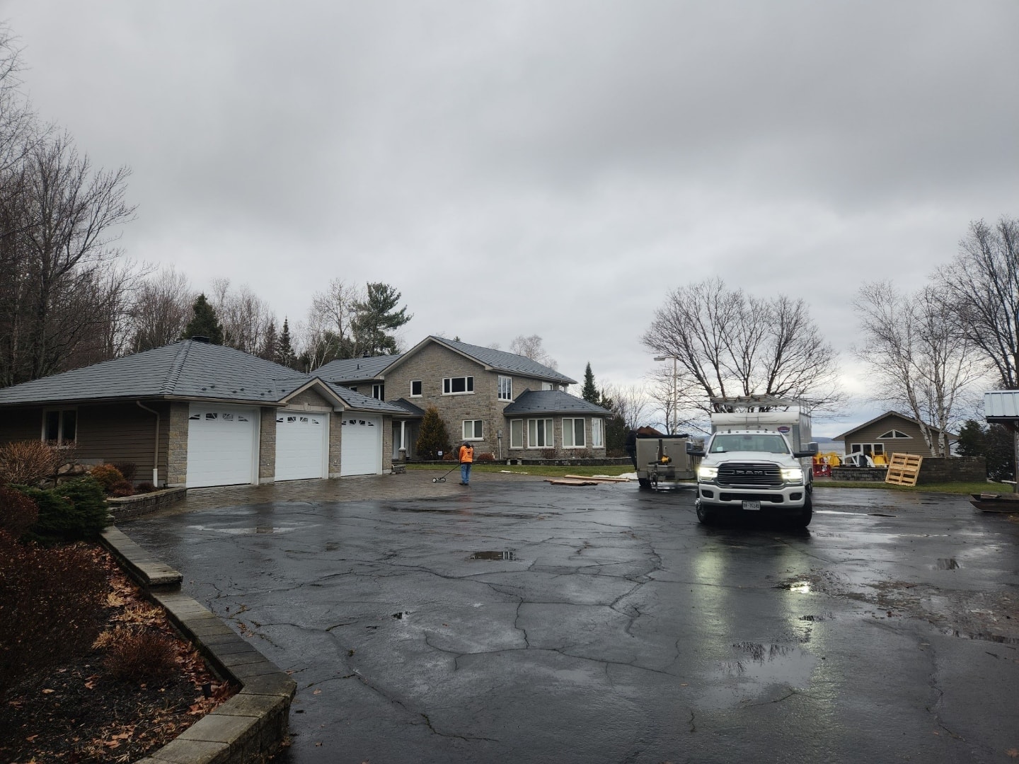 A large suburban house and a three-car garage with metal roofing sits in a spacious driveway. Two workers in orange vests are in the driveway, with one truck and scattered equipment. 