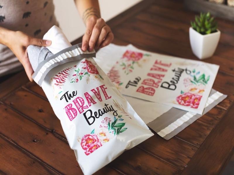 A person placing clothing items into a branded shipping bag with the words "The Brave Beauty" written on it, positioned on a wooden table with a small plant, showcasing a stylish and organized packaging process for a fashion or beauty brand.