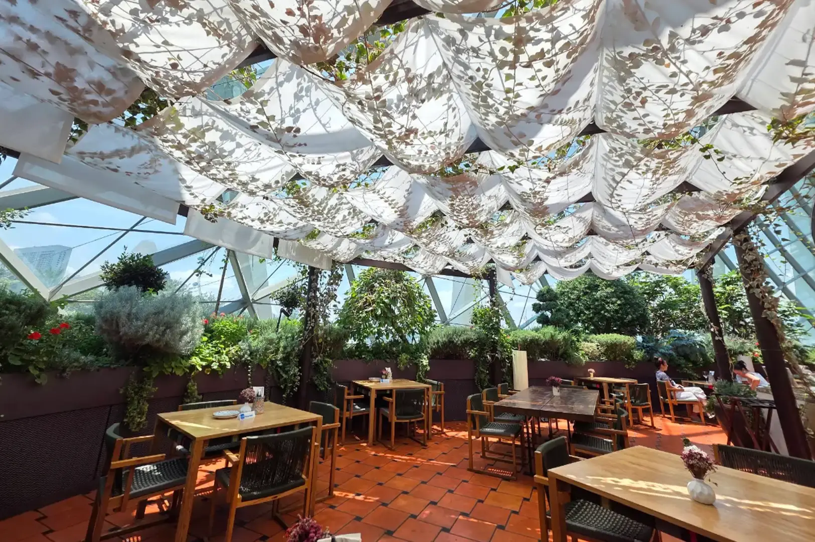 An indoor-outdoor restaurant dining room beneath a glass dome structure, filled with lush garden foliage and wood tables with woven chairs over terracotta tiles, shaded by soft, draped white fabric suspended across the ceiling.