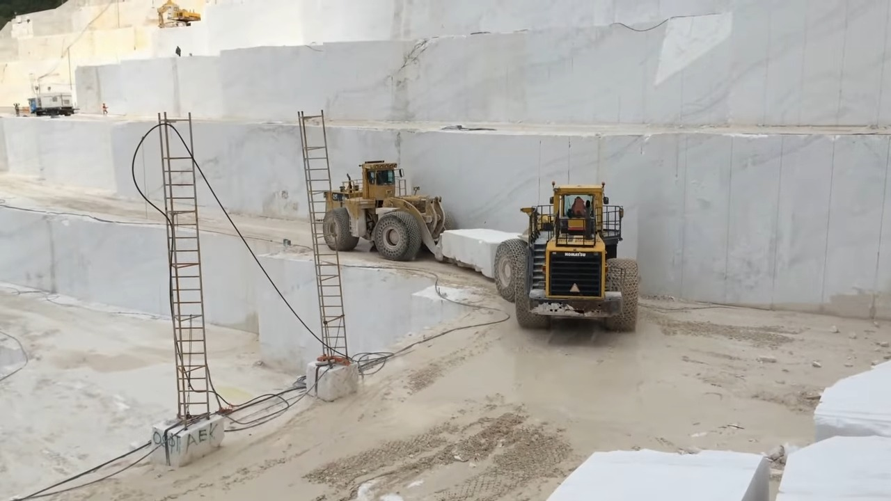 Two large yellow bulldozers with big wheels hauling massive blocks of white marble in a quarry with white marble walls in the background