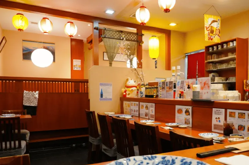 A brightly lit, casual Japanese izakaya interior decorated with warm paper lanterns, featuring wooden counter seating and dining tables prepared for customers.