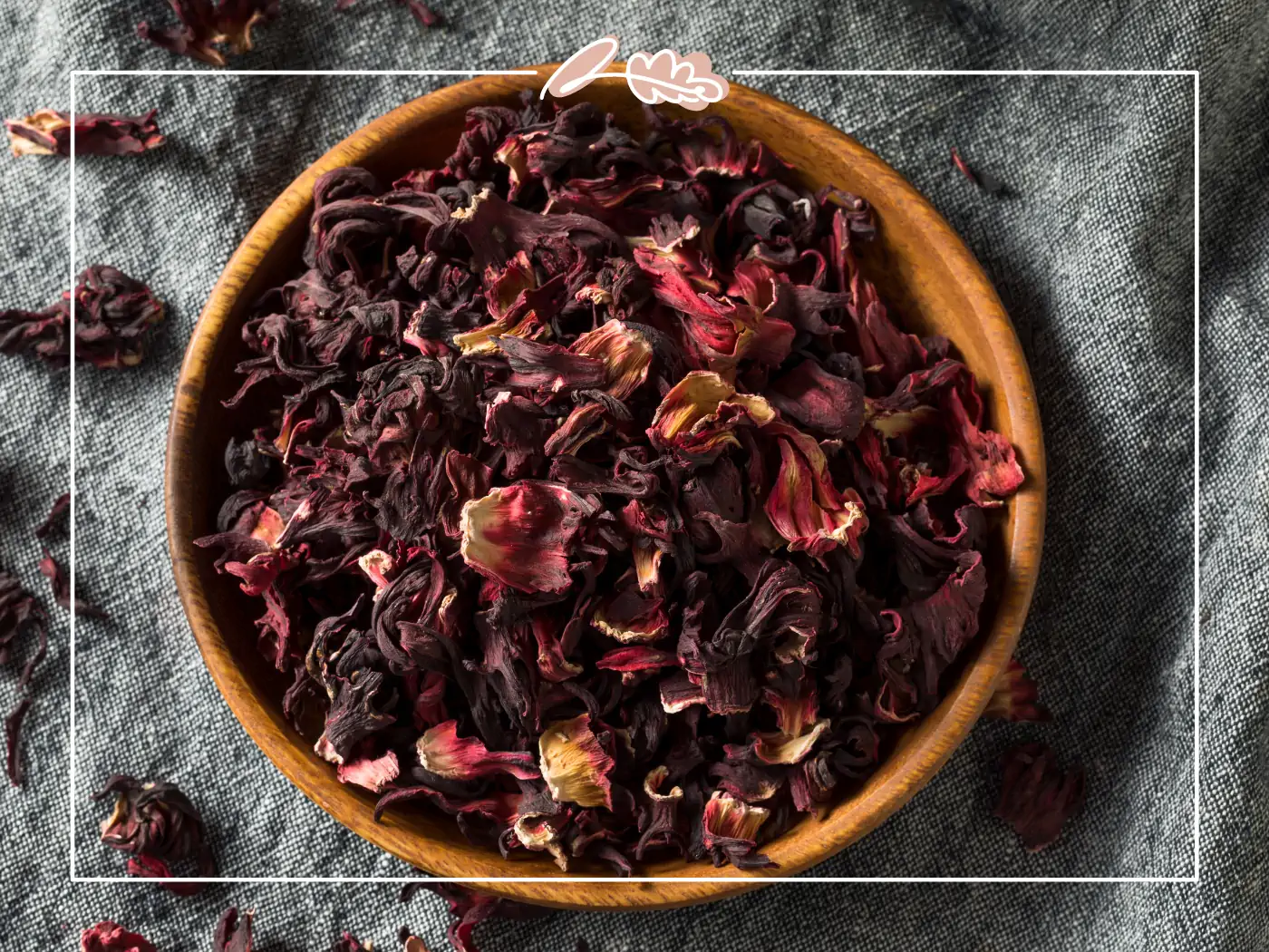 A wooden bowl filled with dried hibiscus petals in rich shades of burgundy and red, placed on a textured grey fabric. Fabulous Flowers and Gifts.