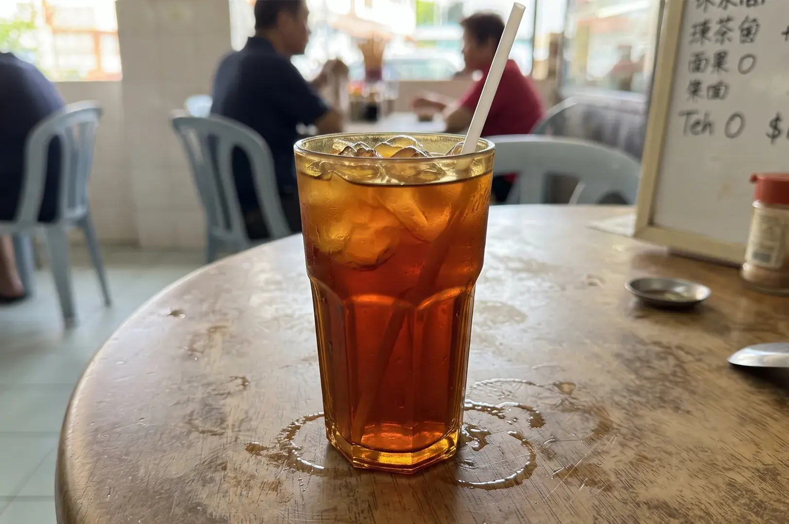 Eye-level close-up of a tall glass of iced black tea with a straw sitting on a wooden dining table inside a local coffeeshop.