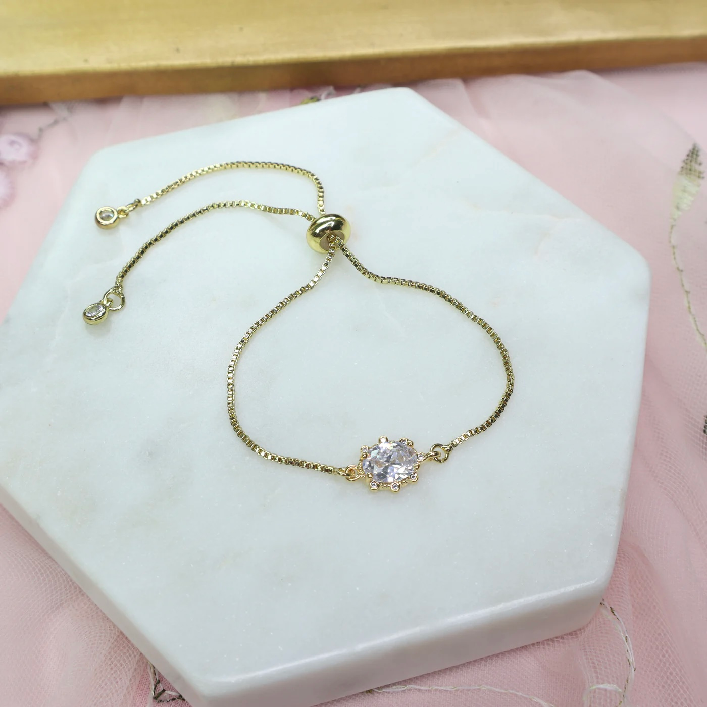 A dainty bracelet with a thin, gold chain and a clear, shiny zircon in the center, laid on a small white platform against a pink cloth.