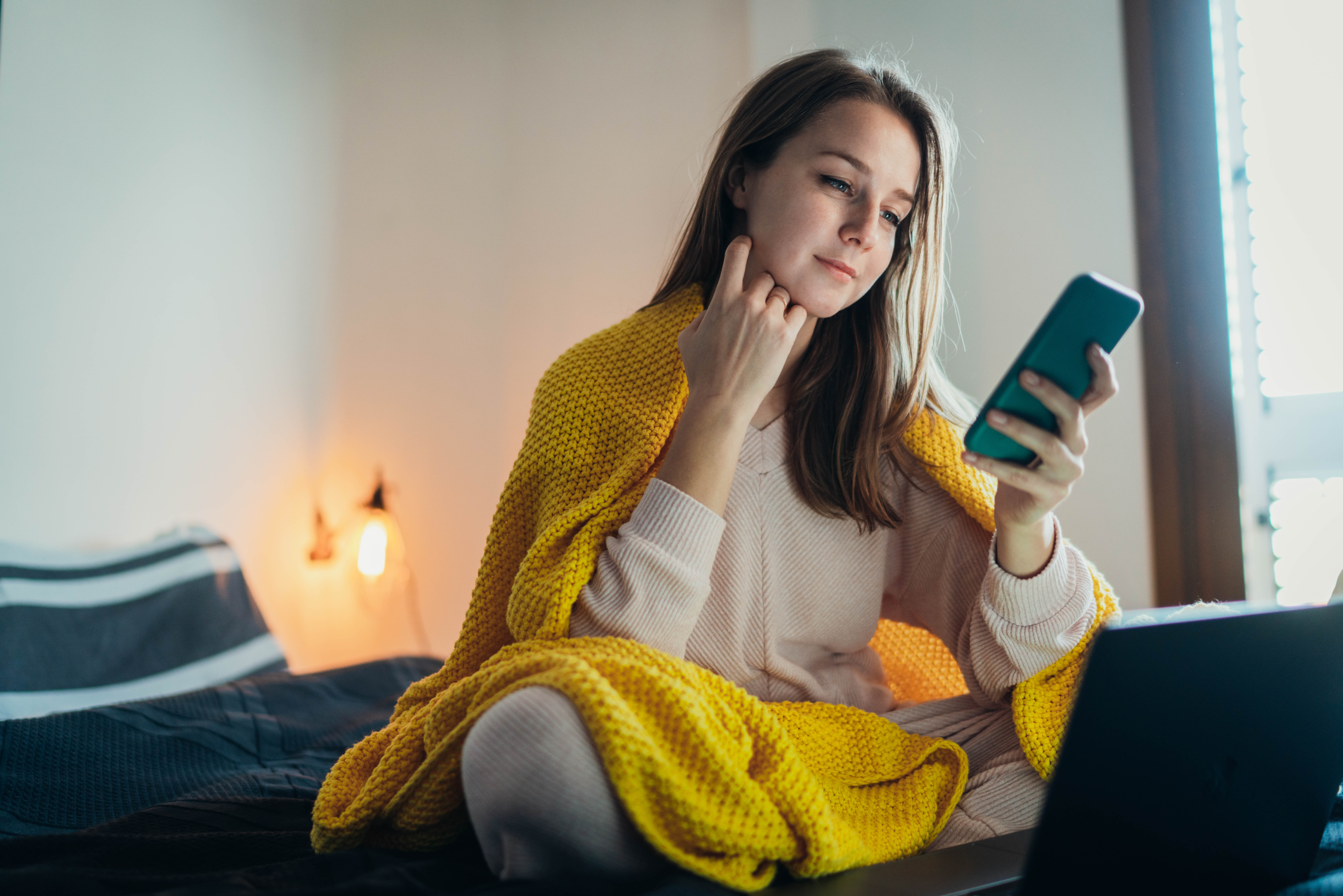 a woman sits on her bed wrapped in a yellow blanket looking at her phone