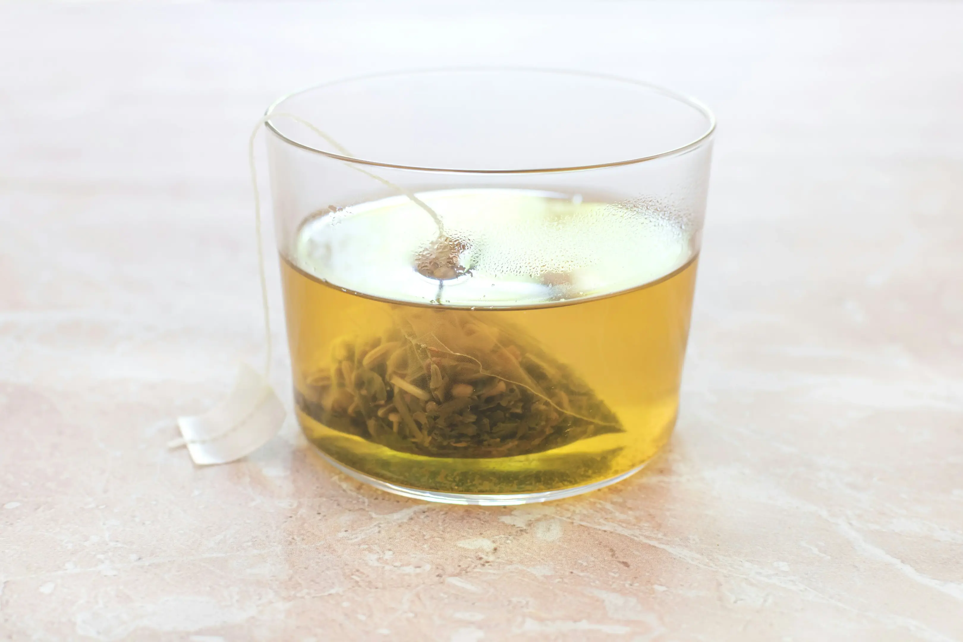 A clear glass cup sits on a marbled pink surface, filled with a light amber-colored tea. Inside the cup, a mesh pyramid tea bag is steeping, with its white string and tag hanging over the side.