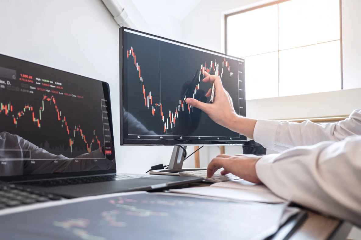 A person points to a stock market chart on a monitor while typing on a keyboard, with another monitor and laptop displaying similar charts. This reflects what is meant by valuation once negotiation begins.