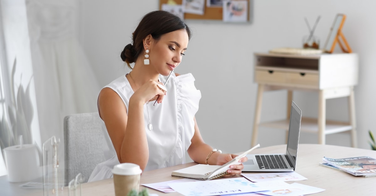 A woman in a white blouse sits at a desk, reviewing documents with a laptop and coffee cup nearby, exemplifying the diligent work of a certified tax planner.