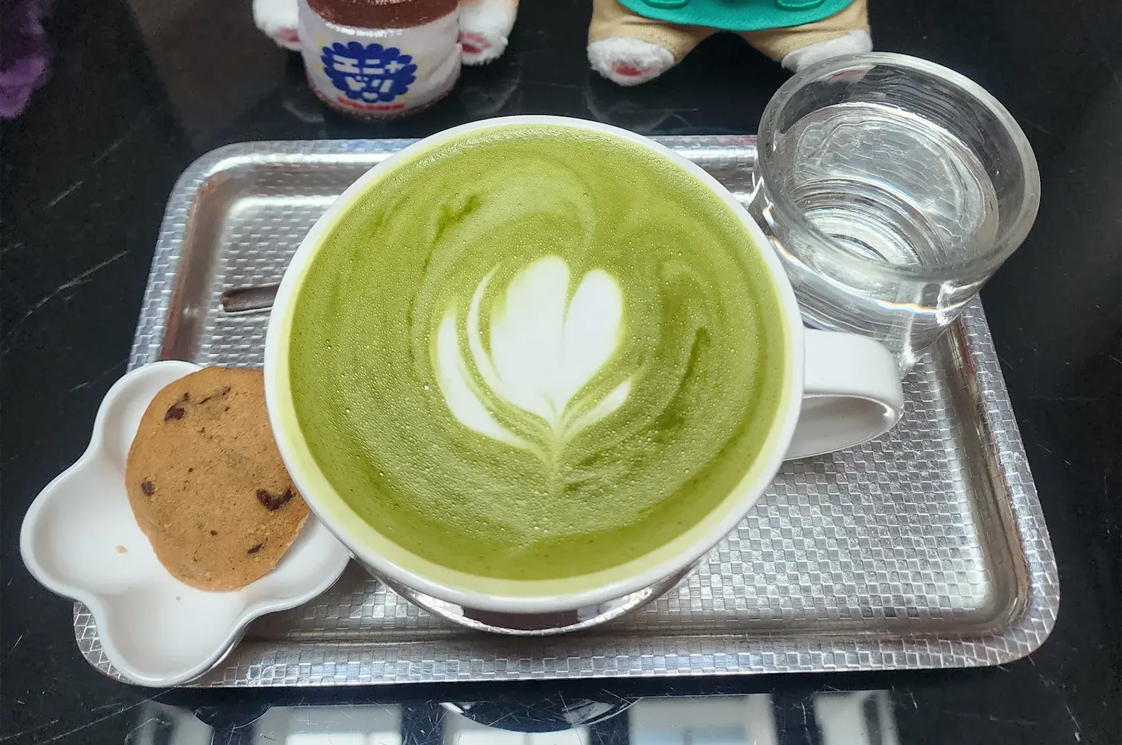 Top-down view of a matcha green tea latte featuring intricate flower leaf latte art, served on a silver tray with a chocolate chip cookie and a glass of water.