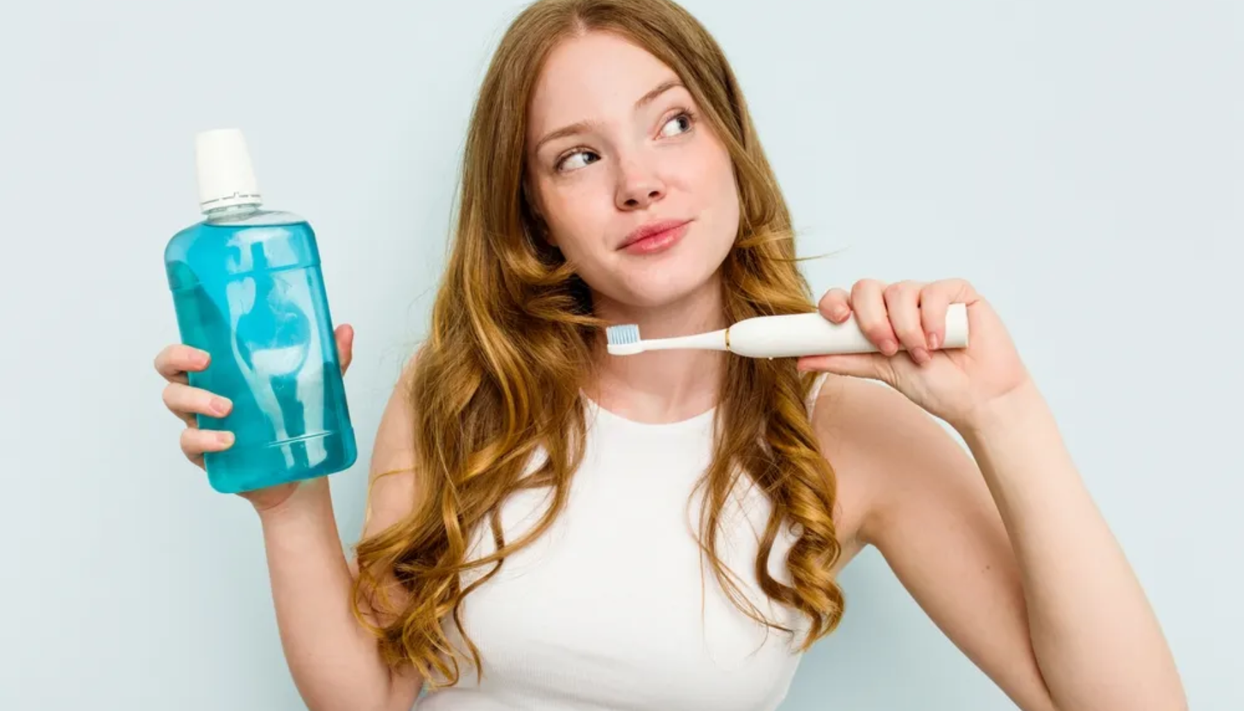 Kvinne holder tannbørste og munnskyll, vurderer munnhygiene med rolig uttrykk mot lys bakgrunn.