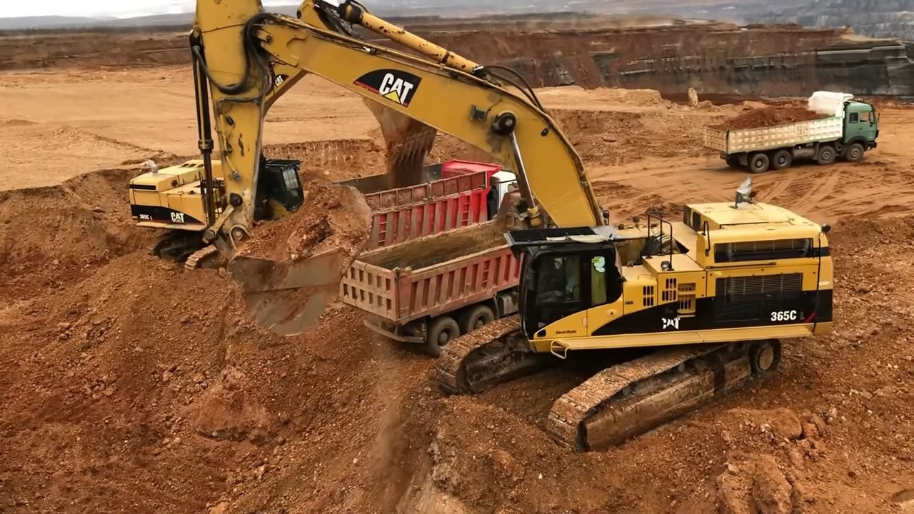 A yellow Caterpillar excavator loading dirt into a red dump truck on a construction site with another truck in the background