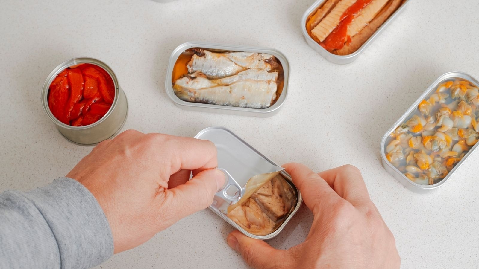 Person opening assorted canned foods including tuna, sardines, and clams on a table