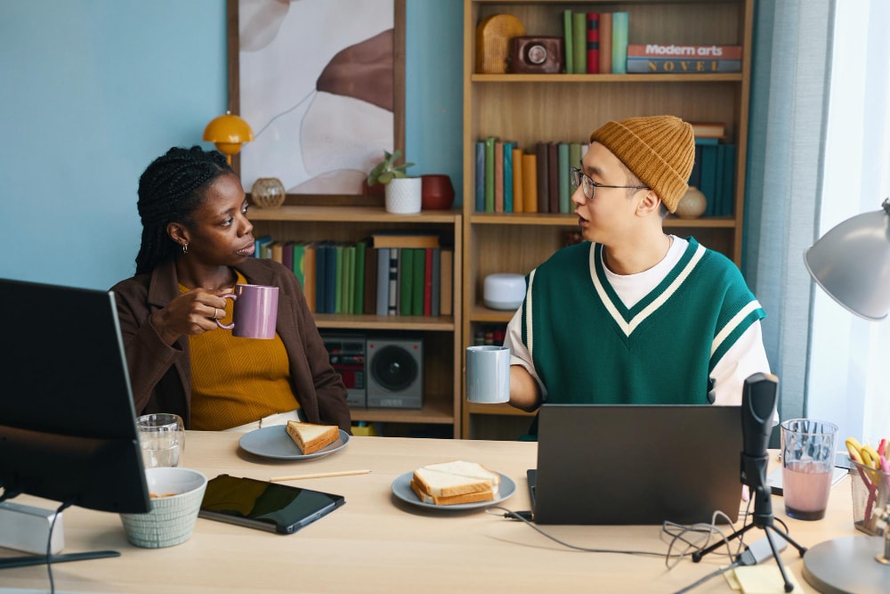 Two coworkers sharing a conversation over coffee at a desk with laptops and sandwiches, illustrating casual yet essential business communication supported by flexible phone system upgrades.