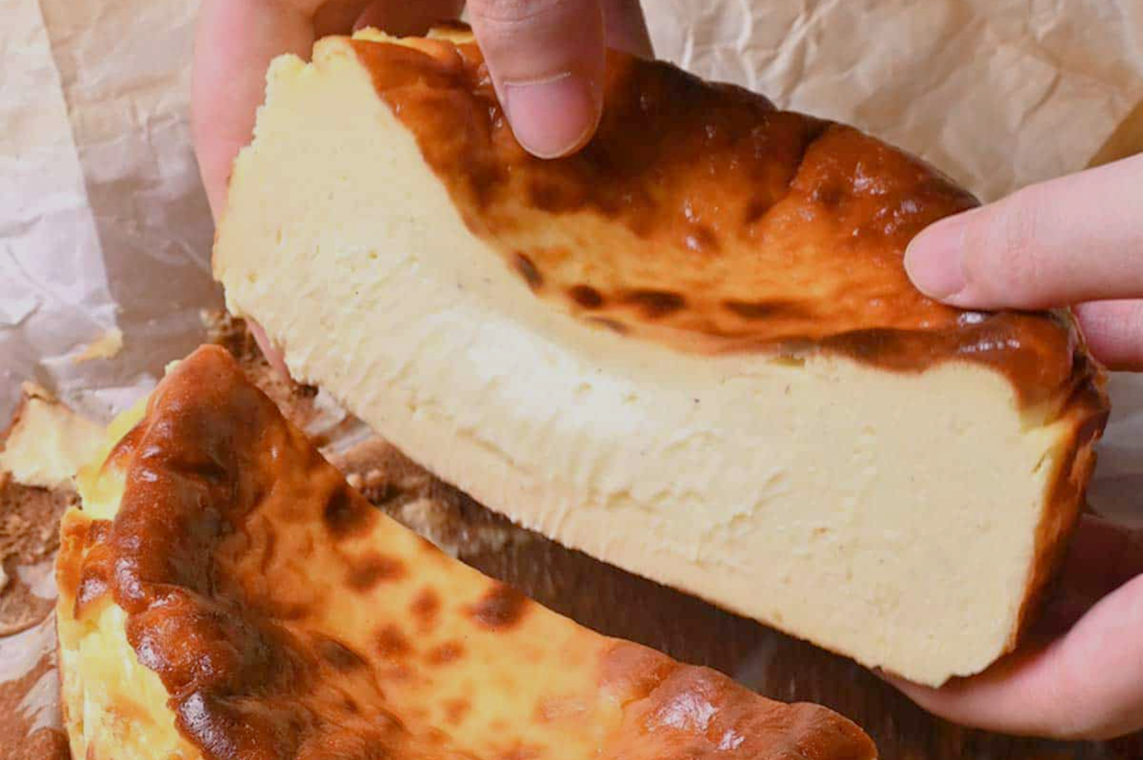 Close-up of hands lifting a slice of creamy Basque burnt cheesecake with a golden-brown top and creamy interior, set against parchment paper.