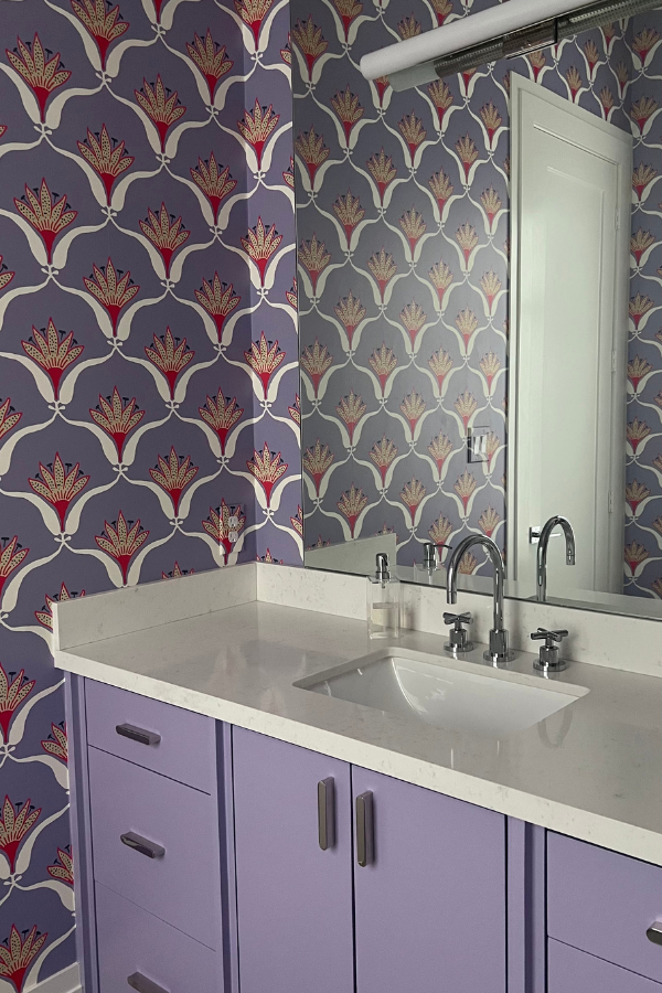 Light purple bathroom vanity with silver hardware and white countertop set against geometric floral wallpaper in purple, red, and gray tones.