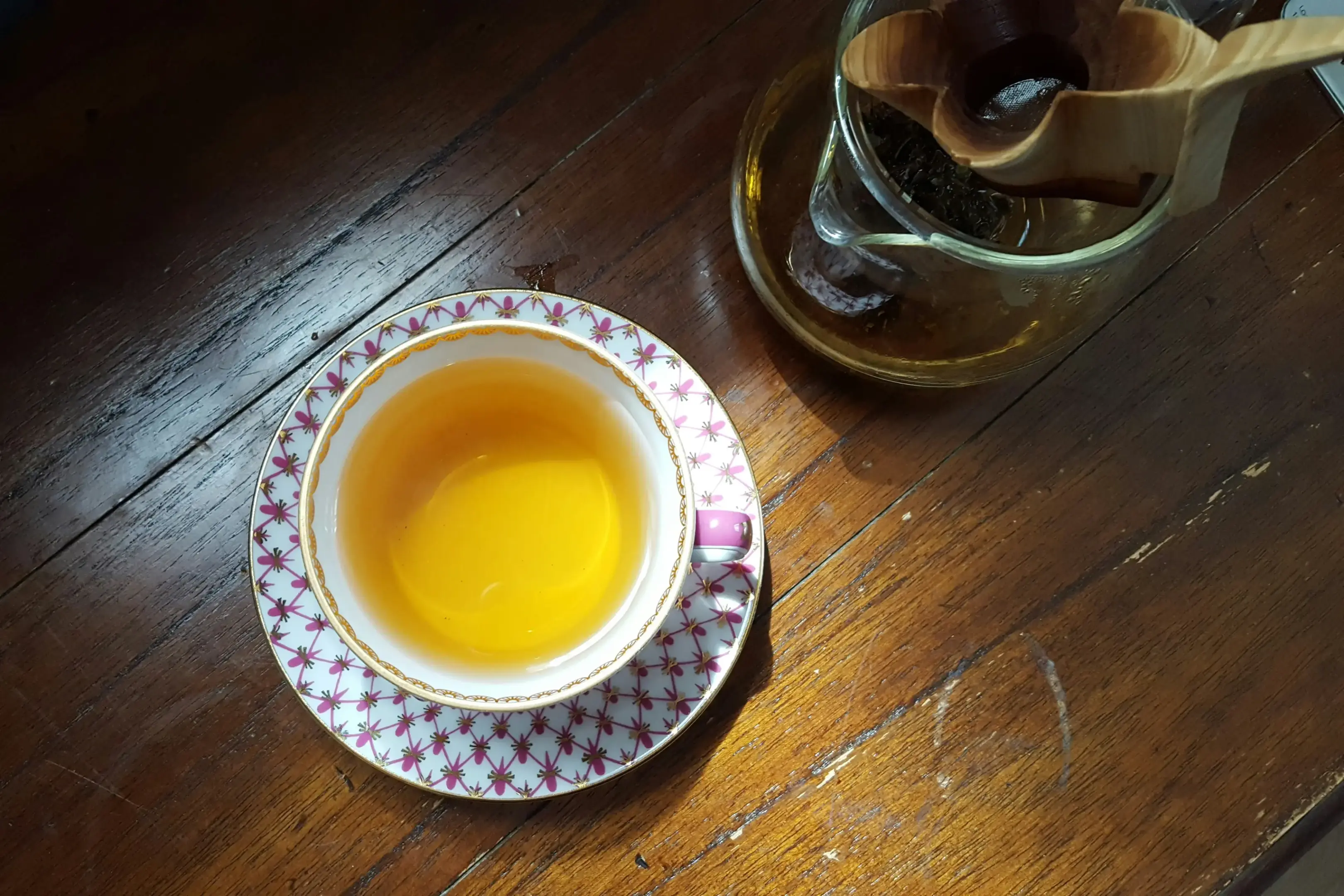 This image captures an overhead view of a delicate porcelain teacup and saucer filled with golden tea, resting on a rustic wooden table. Beside the cup sits a glass carafe featuring a unique wooden flower-shaped tea strainer.