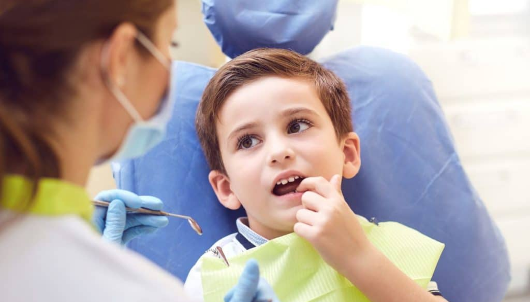 Gutt sitter i tannlegestol og peker på en tann mens tannlegen undersøker.
