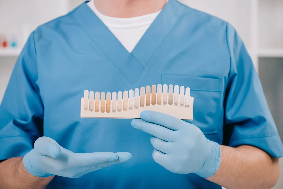 Dental professional in blue scrubs holding a tooth shade guide with various color samples ranging from light to dark, wearing blue examination gloves in a clinical setting. Retry