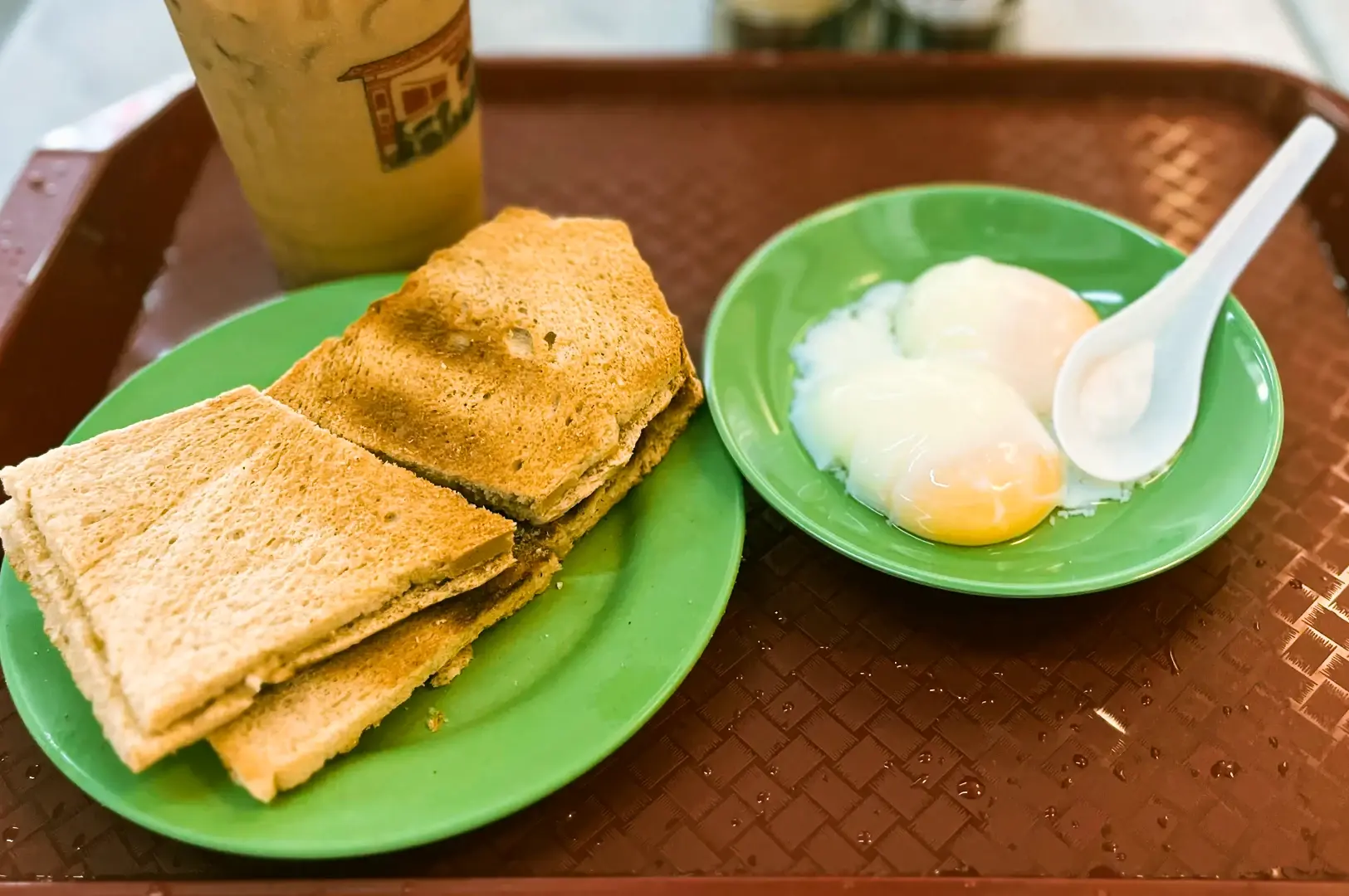 Eye‑level close‑up shot of a traditional Singapore breakfast set featuring kaya toast stacked on a green plate, soft‑boiled eggs in a shallow bowl with a spoon, and a cold drink on a tray at a hawker centre table.