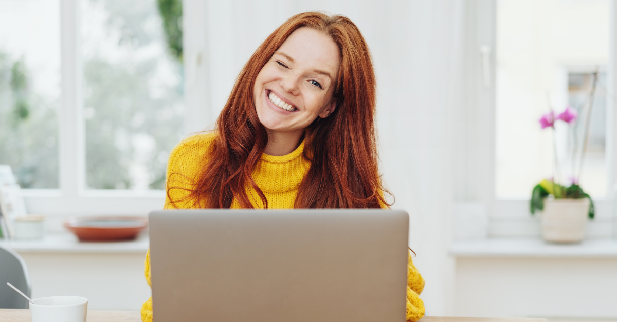 A cheerful red-haired woman winking at her laptop; researching tax and compliance requirements online.