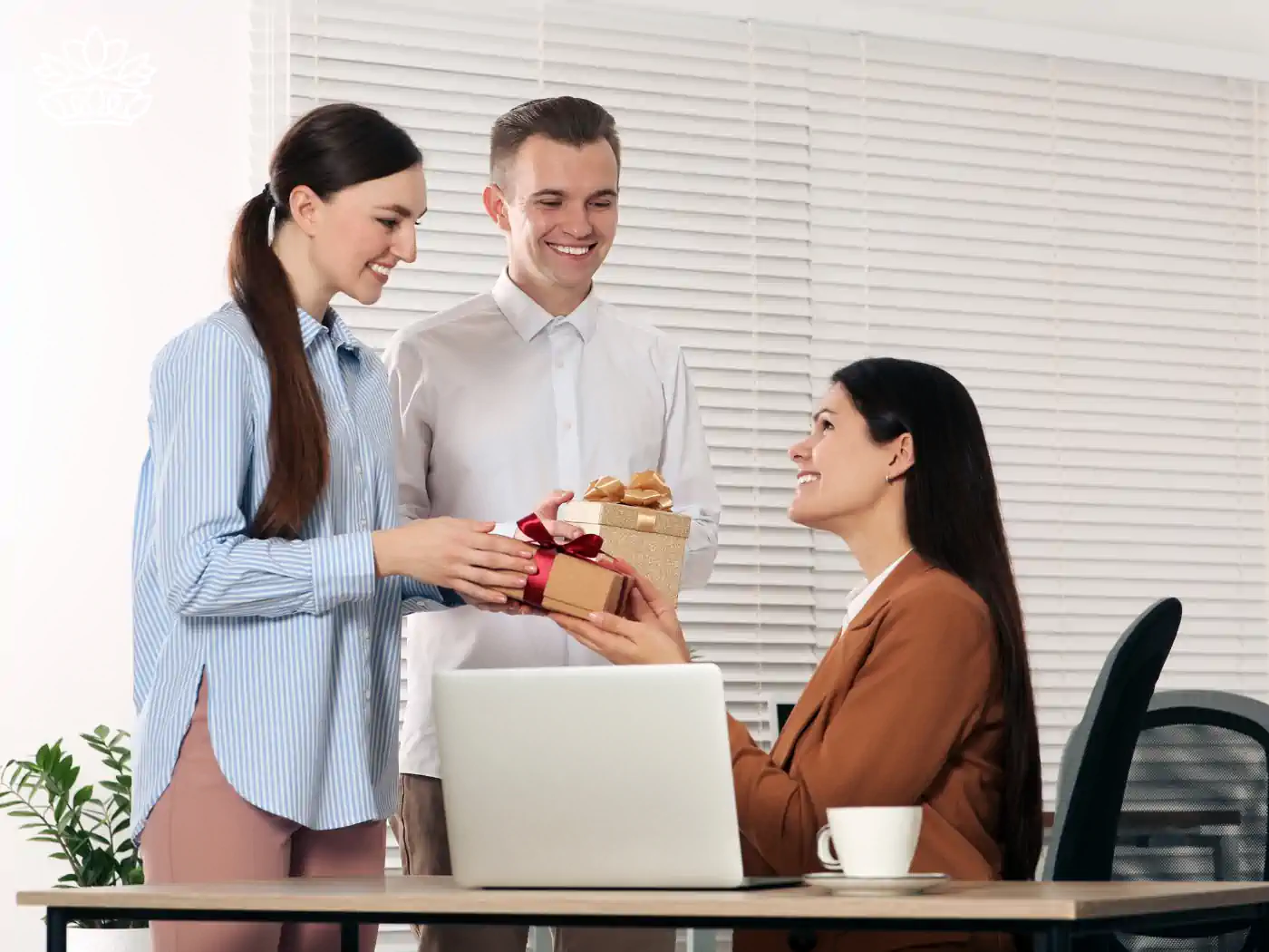 Two colleagues presenting a gift box with a red ribbon to a smiling seated woman in an office, creating a moment of joy and appreciation. The setting conveys a professional yet warm atmosphere, perfect for fostering connections. Gift boxes for colleagues, delivered with heart by Fabulous Flowers and Gifts.
