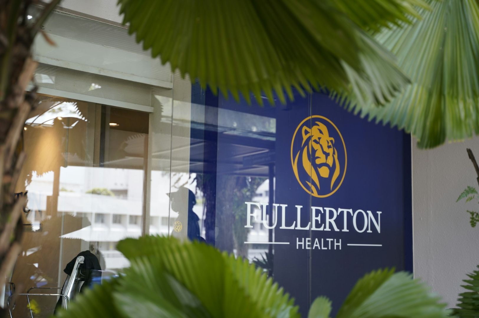 A blue sign with a lion logo and the text "Fullerton Health" is partially obscured by large green leaves, conveying a sense of calm and privacy.