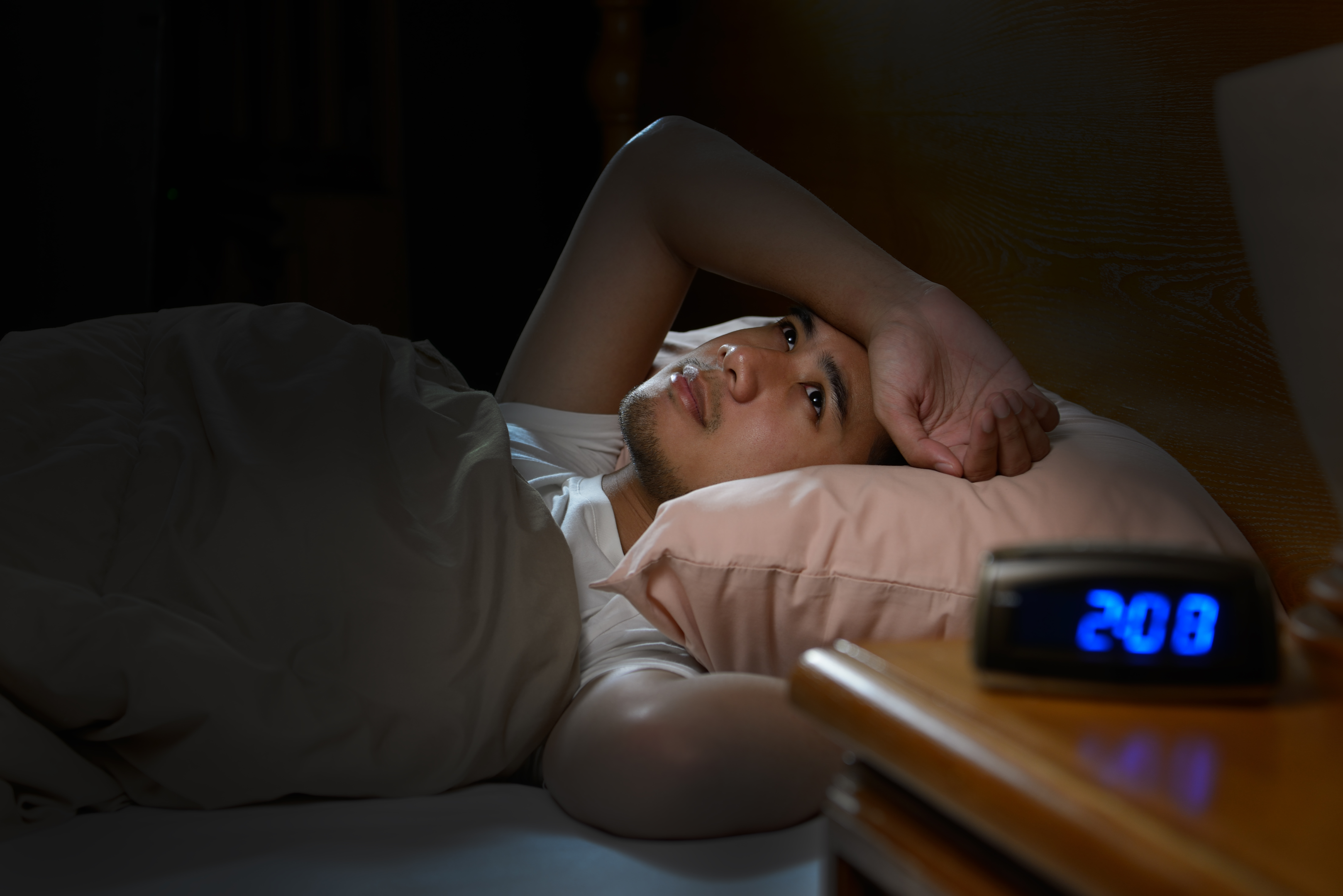 Image of an Asian man laying in bed, looking at the ceiling with one arm resting on his forehead. In the foreground there is a picture of an alarm clock reading 2:00